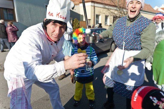 Ein Mann in einer Kochmütze balanciert ein Ei auf einem Löffel, während eine Frau in einem gepunkteten Kleid einen Schüssel hält. Ein Kind in Clownskostüm beobachtet in der Nähe.