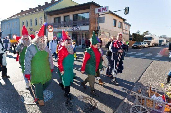 Eine Gruppe von Menschen in Kostümen geht während einer Parade die Straße entlang. Sie tragen rote und grüne Mützen und Schürzen. Im Hintergrund befindet sich ein Gebäude mit einem Schild, das BIPA steht.