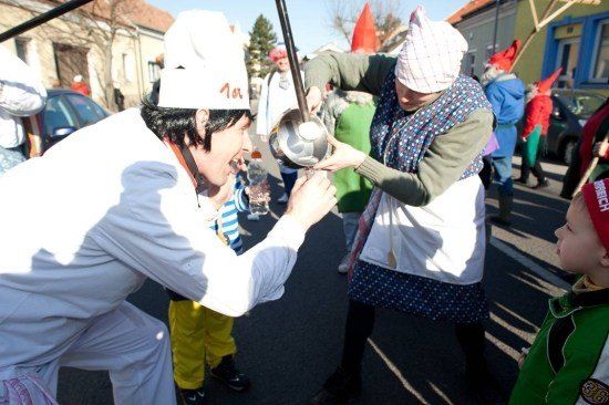 Ein Mann in einer Kochmütze gießt etwas in die Schüssel einer Frau während eines Straßenfestes mit Menschen in Kostümen und einem Kind, das zusieht.