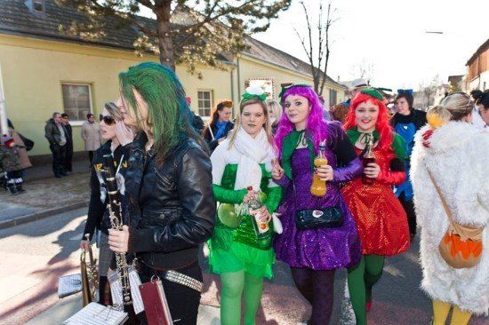Eine Gruppe von Menschen in bunten Kostümen geht eine Straße entlang, möglicherweise für einen Umzug. Eine Person hält ein Musikinstrument.