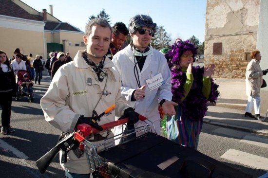 Zwei Männer in Kostümen, einer hält einen Einkaufswagen mit einer angefügten Waffe, gehen in einer Parade mit anderen in Kostümen und einem Kinderwagen im Hintergrund.