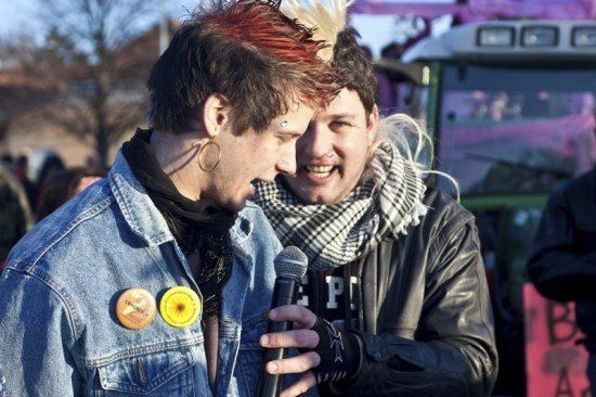 Zwei Männer, einer mit roten Haaren und einer Jeansjacke, spricht in ein Mikrofon, während ein anderer Mann mit einem Schal zusieht. Sie sind in einer Menschenmenge mit einem Fahrzeug im Hintergrund.