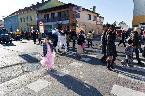 Eine Gruppe von Menschen, darunter ein Kind in einem rosa Kleid, überquert eine Straße in einer Stadt. Einige tragen Kostüme. Im Hintergrund befinden sich Gebäude und ein Schild mit der Aufschrift BIPA.