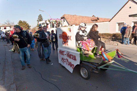 Eine Gruppe von Menschen in Kostümen geht mit einem Wagen die Straße entlang, während andere Musikinstrumente spielen.