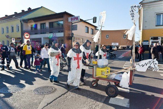 Eine Gruppe von Menschen in mittelalterlichen Ritterkostümen zieht bei einer Parade auf einer Stadtstraße. Sie tragen einen Wagen mit einer Pferdefigur. Hinter ihnen beobachten Menschen die Parade. Im Hintergrund befinden sich Gebäude und Straßenschilder.