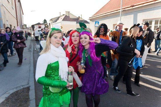 Drei Frauen in Kostümen posieren für ein Foto auf einer Straße während eines Umzugs. Dahinter laufen und stehen Menschen in verschiedenen Kostümen. Gebäude säumen die Straße.