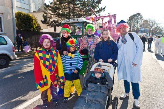 Eine Gruppe von Menschen in Clownkostümen steht auf der Straße, mit einem Baby im Kinderwagen und einem Mann in einem weißen Gewand.