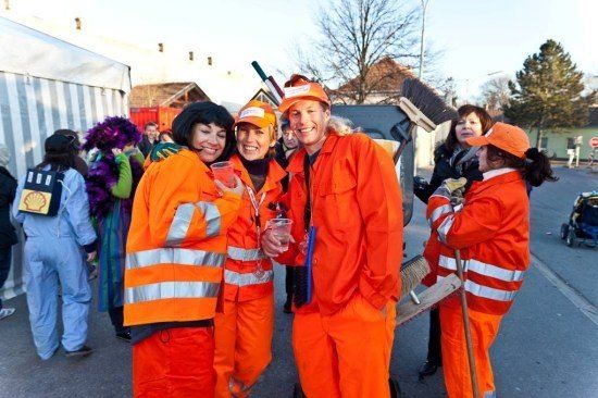 Eine Gruppe von Menschen in orangefarbenen Schutzanzügen und Mützen steht auf der Straße, lächelt und posiert für ein Foto. Eine Frau in der Mitte hält einen Becher. Hinter ihnen reinigt eine Frau die Straße mit einem Besen.