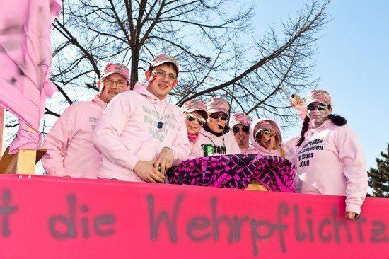 Eine Gruppe von Menschen in rosa Kapuzenpullovern und Hüten posiert für ein Foto auf einem Wagen mit einem rosa Hintergrund und Graffiti. Sie lächeln und scheinen an einer Parade teilzunehmen.