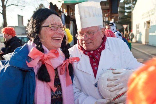 Eine Frau mit Zöpfen und einer rosa Schleife lächelt neben einem Mann in einer Kochmütze, der einen Fußball hält.