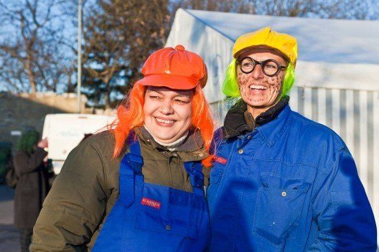 Zwei Personen in farbenfrohen Kostümen, eine mit orangefarbenem Haar und rotem Hut, die andere mit grünem Haar und gelbem Hut, stehen zusammen draußen.