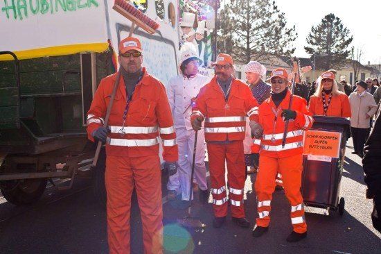 Eine Gruppe von Menschen in orangefarbenen Overalls und Mützen steht vor einem Müllwagen. Dahinter befindet sich eine Umzugswagen mit einem Schild, das 'HAUBINGER' besagt.