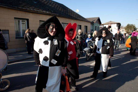 Eine Gruppe von Menschen in Kostümen geht die Straße entlang, möglicherweise für einen Umzug. Sie tragen Schminke im Gesicht und halten Taschen. Hinter ihnen stehen Gebäude, und einige Leute stehen und beobachten.
