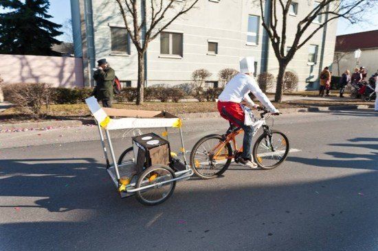 Eine Person fährt mit einem an den Hinterreifen angeschlossenen Kasten auf einem Fahrrad und trägt eine Kochmütze. Ein Mann steht in der Nähe und Menschen gehen auf dem Bürgersteig.