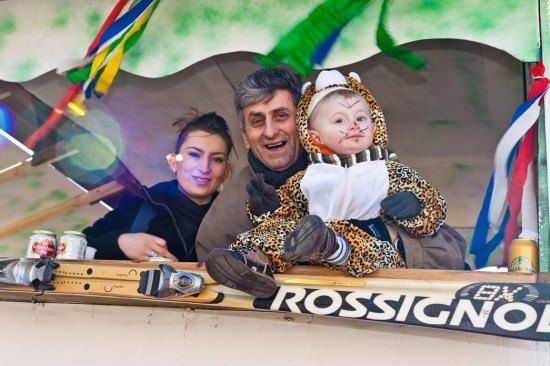 Eine dreiköpfige Familie, bestehend aus einem Mann, einer Frau und einem Baby in einem Leopardenkostüm, lächelt und posiert für ein Foto in einem dekorierten Stand mit einem Schild, das 'Rossignol' liest.