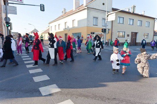 Eine Gruppe von Menschen in verschiedenen Kostümen geht auf der Straße, wahrscheinlich für einen Umzug. Einige tragen Hüte und manche haben Taschen. Die Straße hat weiße Markierungen und Ampeln. Gebäude mit Fenstern befinden sich auf beiden Seiten der Straße.