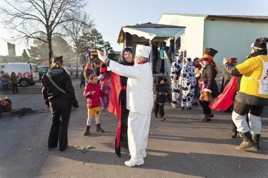 Eine Gruppe von Menschen in Kostümen, darunter ein Koch, tanzt auf einem Freigelände mit einem Gebäude und geparkten Fahrzeugen im Hintergrund.