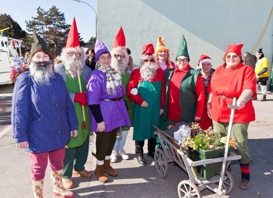 Eine Gruppe von Menschen, die als Elfen gekleidet sind, einige in Kostümen und andere in festlicher Kleidung, stehen vor einem Gebäude mit einem Blumenwagen.