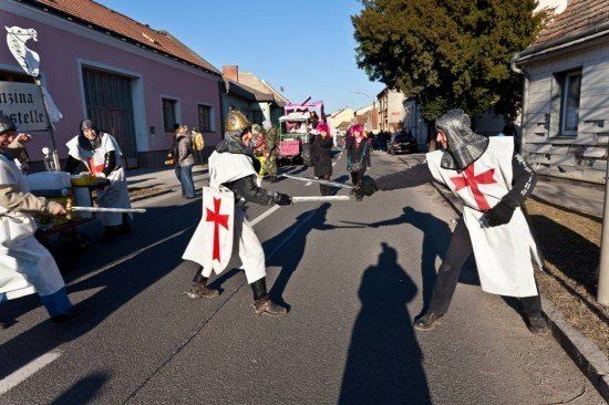 Mehrere Personen, die als Ritter verkleidet sind, führen eine Scheinschwertkampf in der Mitte einer Straße. Zuschauer beobachten von den Seitenlinien.