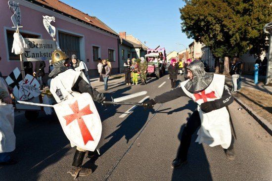 Zwei Ritter in mittelalterlichen Kostümen führen eine Scheinschwertkampf in der Mitte einer Straße, mit Zuschauern und einem rosafarbenen Wagen im Hintergrund.