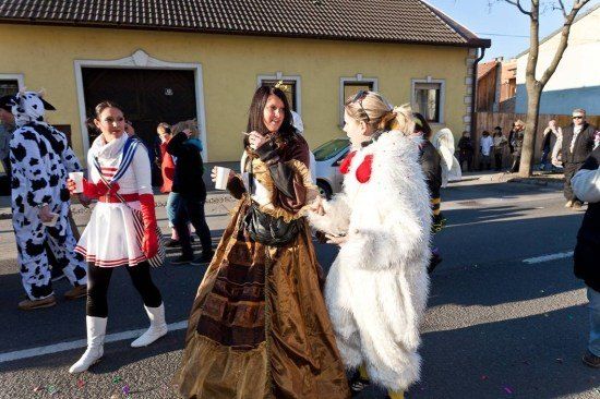 Drei Frauen in Kostümen gehen eine Straße entlang, eine in einem Kuhkostüm, eine andere in einem mittelalterlichen Kleid und die dritte in einem weißen Pelzengel-Outfit. Hinter ihnen gehen und stehen weitere Menschen in Kostümen in der Nähe eines gelben Gebäudes.