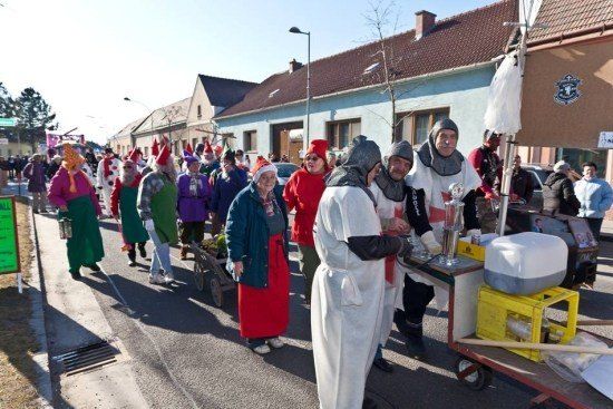Eine Gruppe von Menschen in bunten Kostümen geht auf einer Straße, einige tragen Hüte und Brillen. Ein Mann in einem weißen Gewand steht neben einem Wagen mit einer Kanne.