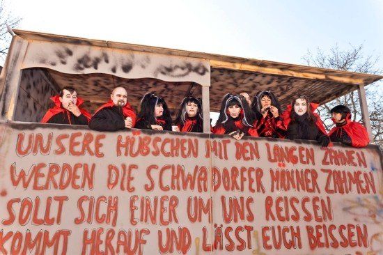 Eine Gruppe von Menschen in roten Kostümen steht auf einer Bühne mit einem Banner, auf dem steht 'Unsere Hübschen mit den langen Zähnen werden die schwarzen Dörfer Männer zähmen'. Sie scheinen eine Aufführung zu machen oder an einer Parade teilzunehmen.