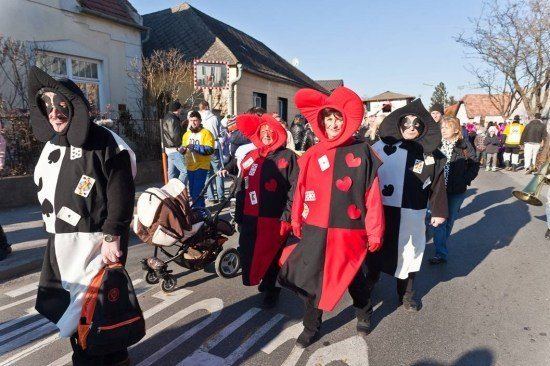 Eine Gruppe von Menschen in Kostümen geht während einer Parade die Straße entlang, einer hält einen Rucksack und ein anderer schiebt einen Kinderwagen.