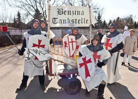 Eine Gruppe von Menschen, die als Ritter mit roten Kreuzen auf ihren Schilden gekleidet sind, posiert für ein Foto vor einem Wagen mit einem Schild, das Benzina Tankstelle liest.