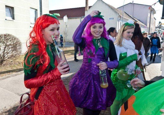 Drei Frauen in bunten Kostümen, möglicherweise für ein Fest, stehen auf einer Straße mit Flaschen und einem grünen Apfel. Eine Frau hat rote Haare und ein rotes Kleid, eine andere hat lila Haare und ein lila Kleid und die dritte hat blonde Haare und ein grünes Kleid.