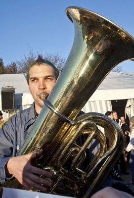 Ein Mann mit grünen Haaren spielt ein großes Messing-Tuba bei einer Veranstaltung im Freien, mit einem verschwommenen Publikum im Hintergrund.