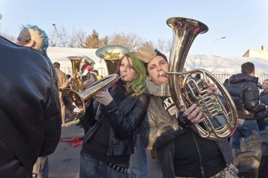 Zwei Personen mit grünen Haaren spielen Blechblasinstrumente in einer Freiluftumgebung, umgeben von anderen Musikern und Zuschauern.