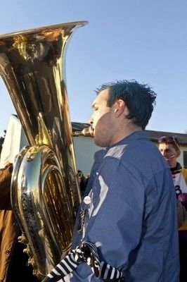 Ein Mann mit grünen Haaren spielt ein Messing-Tuba im Freien, mit einer weiteren Person in einem gelben und schwarzen Trikot im Hintergrund.
