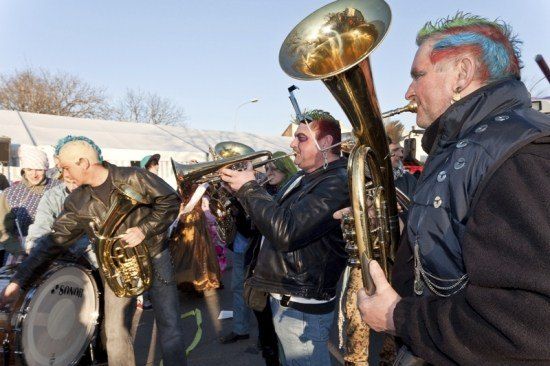 Eine Gruppe von Musikern in Lederjacken und Gesichtsbemalung spielt Blechblasinstrumente im Freien, während eine Menge zuschaut.