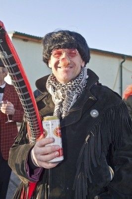Ein Mann in einer Jacke mit Fransen und einem Schal hält eine Dose Bier und ein langes, schwarzes Objekt, möglicherweise ein Musikinstrument, während er draußen steht.