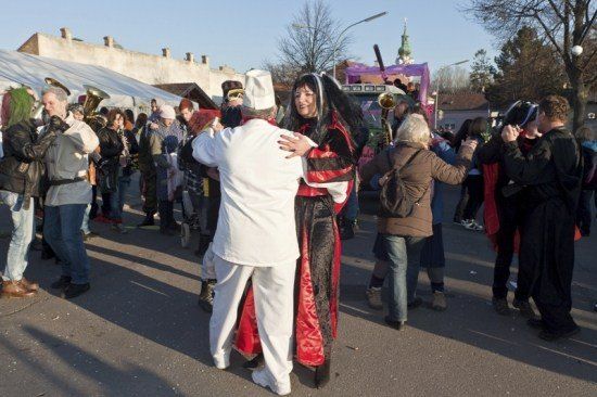 Eine Gruppe von Menschen in Kostümen, die auf einem Freigelände tanzen, wahrscheinlich bei einem Festival. Ein Mann in einer Kochmütze und eine Frau in einem roten und schwarzen Kostüm tanzen zusammen.