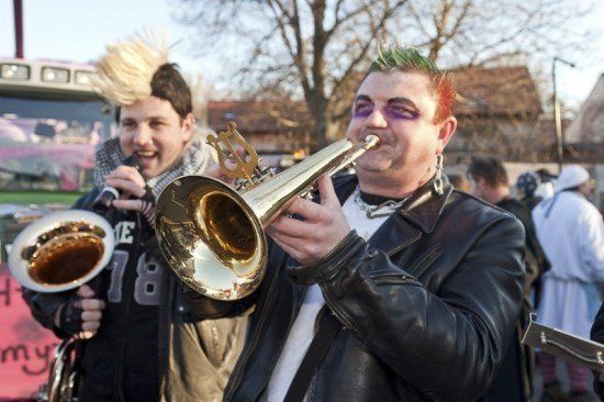 Zwei Männer in Punk-Kleidung spielen Posaunen in einer Outdoor-Umgebung mit einer Menschenmenge im Hintergrund.