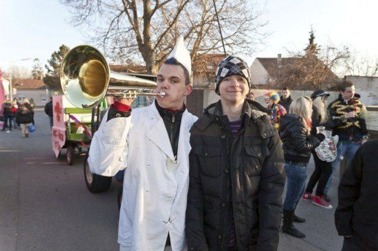 Zwei Männer stehen zusammen auf einer Straße bei einem festlichen Ereignis, einer spielt Posaune und der andere lächelt. Dahinter sind Menschen in bunten Kostümen und ein geschmückter Wagen zu sehen.