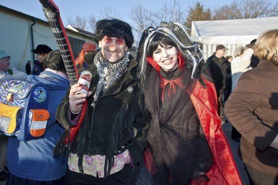 Zwei Menschen in Kostümen posieren für ein Foto bei einer Outdoor-Veranstaltung. Der Mann hält eine Gitarre und eine Dose. Sie sind von anderen Menschen umgeben, von denen einige ebenfalls kostümiert sind.