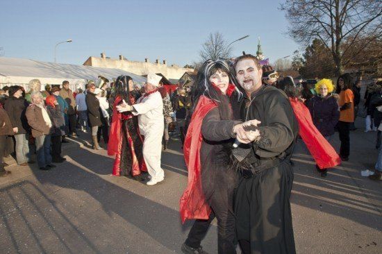 Eine Gruppe von Menschen in verschiedenen Kostümen, darunter ein Vampir und ein Koch, laufen in einem Umzug auf einer Straße.