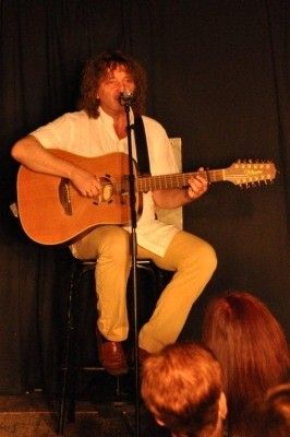 Ein Mann mit lockigen Haaren sitzt auf einem Hocker und spielt Gitarre vor einem Mikrofon, während er auf der Bühne auftritt.