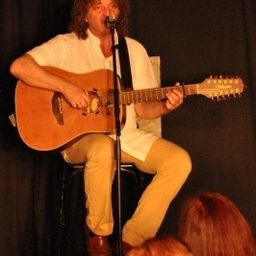 Ein Mann mit lockigen Haaren sitzt auf einem Hocker und spielt Gitarre vor einem Mikrofon, während er auf der Bühne auftritt.