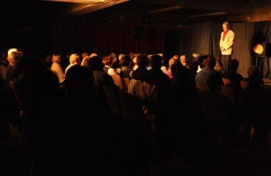 Ein Mann steht auf der Bühne in einem schwach beleuchteten Raum und spricht ein Publikum an, das auf Stühlen sitzt. Die Bühne ist von einem Scheinwerfer beleuchtet.
