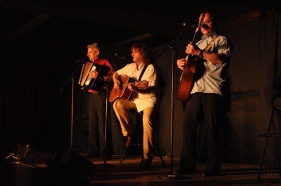 Drei Männer spielen auf der Bühne mit Gitarren und einem Akkordeon, beleuchtet von einem Scheinwerfer.