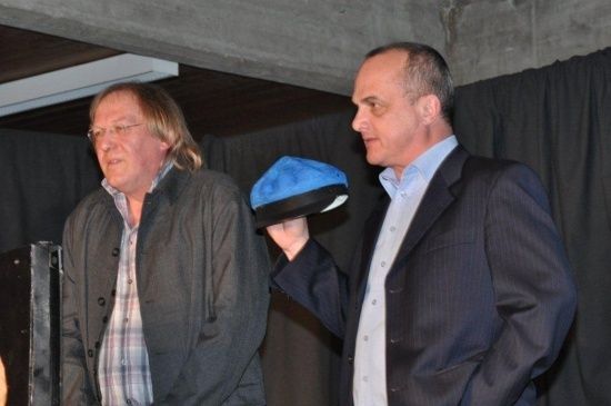 Zwei Männer stehen nebeneinander, einer hält einen blauen Hut, beide sind formell gekleidet, möglicherweise bei einer Veranstaltung.