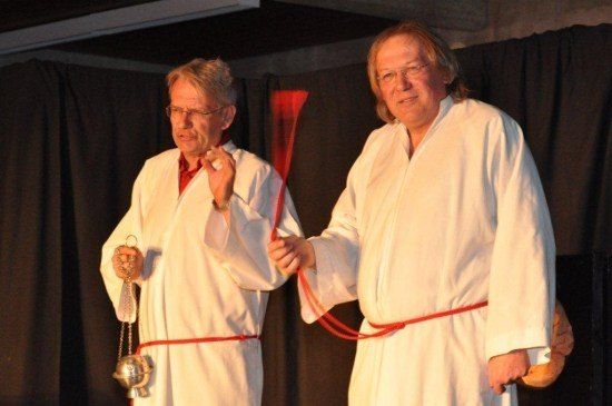 Zwei Männer in weißen Gewändern, einer hält eine Glocke und der andere ein rotes Band, stehen auf der Bühne.