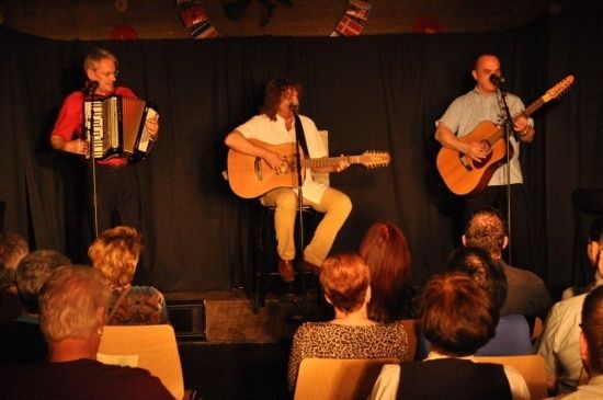 Drei Musiker spielen auf der Bühne vor einem Publikum im Hintergrund. Einer spielt Akkordeon, ein anderer spielt Gitarre und singt, und der dritte spielt Gitarre und singt.