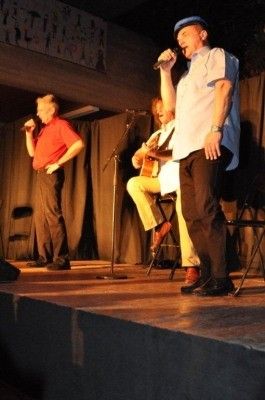 Drei Männer auf der Bühne, einer singt in ein Mikrofon, ein anderer spielt Gitarre, der dritte sitzt mit einer Gitarre, alle unter einem Scheinwerfer.