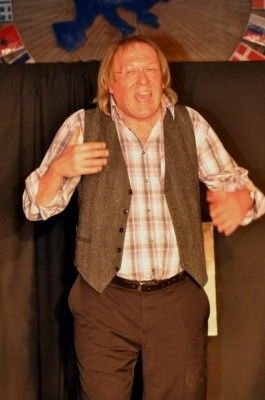 Ein Mann mit langen blonden Haaren und einem Bart steht auf der Bühne, trägt ein kariertes Hemd, eine Weste und braune Hosen. Er macht eine Handgeste mit der rechten Hand.