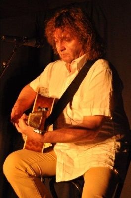 Ein Mann mit lockigem Haar sitzt auf einem Stuhl und spielt Gitarre. Er trägt ein weißes Hemd und beige Hose. Ein Mikrofon steht vor ihm.